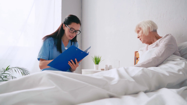 Yatakta yaşlı bir kadın, panoya yazı yazan yaşlı bir doktorla konuşuyor. - Video, Çekim