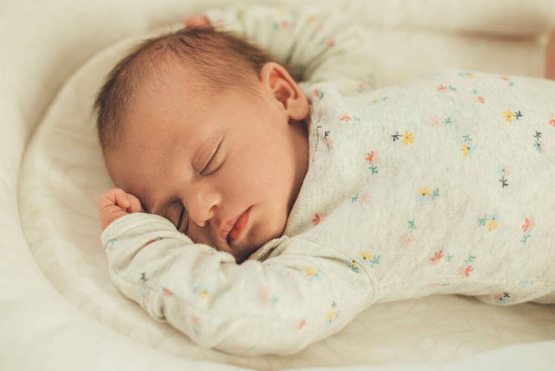 Новорожденный ребенок в белой теплой одежде спит в постели полностью расслабленным и чувствовать себя в безопасности - Фото, изображение