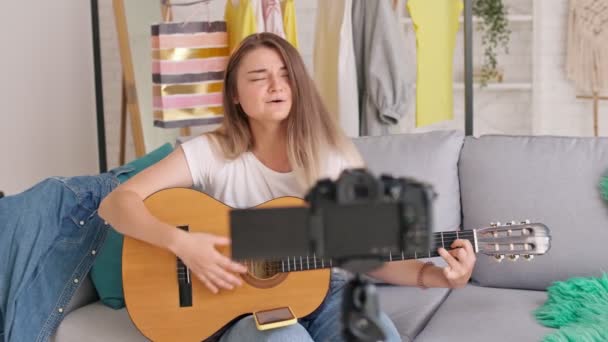 Ein attraktives junges Mädchen bringt beim Bloggen aus der Ferne das Gitarrespielen bei. Mädchen spricht während der Dreharbeiten zu ihrem Videoblog im Hellen Raum - Filmmaterial, Video