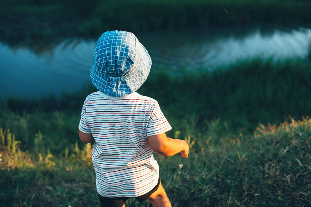 Caucasian boy looking at a small river and throwing stones wearing a blue hat in a green field - Foto, Bild