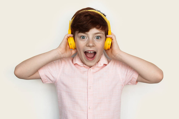 Amazed caucasian ginger boy wearing headphones listening to music on a white studio wall - Foto, Imagen