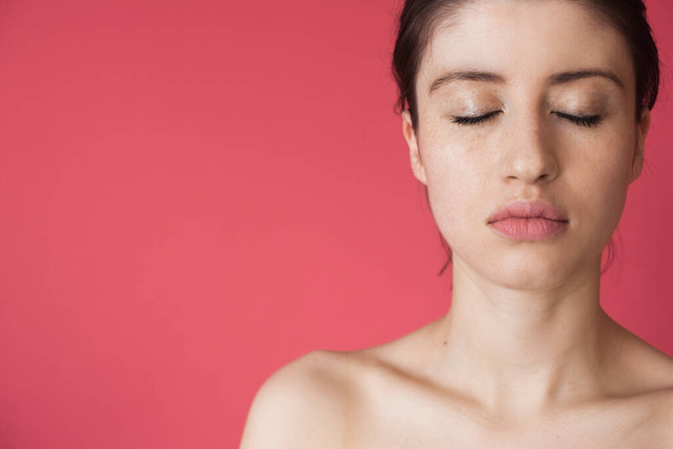 Photo rapprochée d'une femme rousselée avec les yeux fermés posant avec des épaules nues sur un mur rose annonçant quelque chose - Photo, image