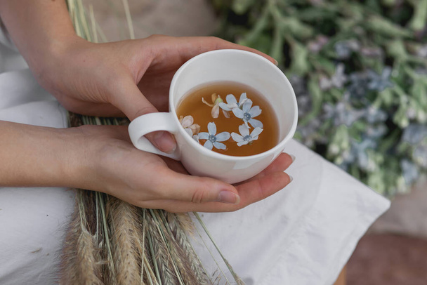 Teetasse in weiblichen Händen eines jungen Mädchens im Freien, mit verschwommenen Blumen auf dem Hintergrund. Becher mit Tee und blauen Blumen innen. - Foto, Bild