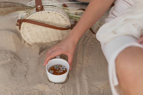 Taza de té en manos femeninas de una joven al aire libre en la playa, con bolsa borrosa con espiguillas en el fondo. Mujer joven en vestido blanco está sosteniendo taza con té y flores en el interior.  - Foto, Imagen