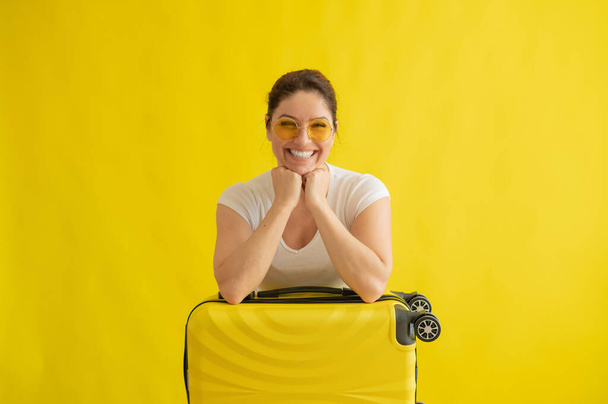 A woman in sunglasses hugs a suitcase on a yellow background - Foto, Bild