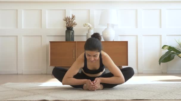 Молодая индийская женщина улыбается медитируя сидя на ковре и делая растяжку, занимаясь йогой, черный спортивная комната дома утром - Кадры, видео