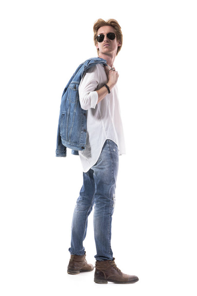 Vista lateral do jovem fotogênico em jeans carregando casaco olhando para trás sobre o ombro. Corpo inteiro isolado em fundo branco. - Foto, Imagem