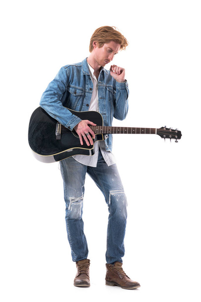 Θλιβερός νεαρός κιθαρίστας που κρατάει ακουστική κιθάρα κοιτώντας κάτω συγκεντρωμένος. Πλήρες σώμα απομονωμένο σε λευκό φόντο. - Φωτογραφία, εικόνα