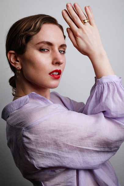 Beautiful young woman with red lips in purple shirt posing with hands up on the white background. - Фото, зображення