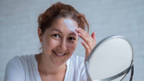Portrait of a Caucasian woman with vitiligo uses sunscreen. A girl with a white pigment spot on her forehead looks in the mirror and is smeared with cream. - Foto, Imagem