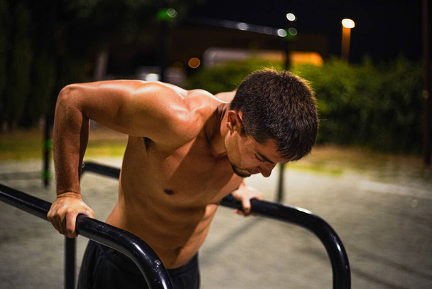 Un joven musculoso sin camisa entrenando en el entrenamiento nocturno en la calle. En un bar público. Haciendo salsas. Fotografía frontal. - Foto, imagen