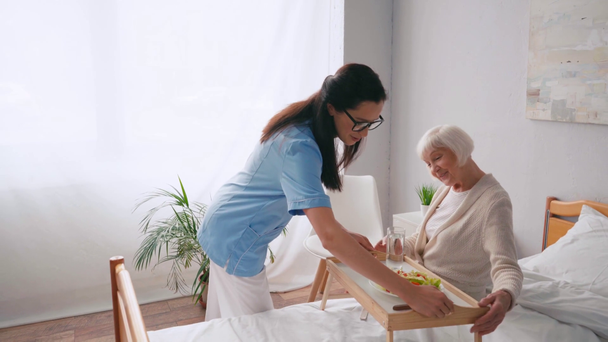 jovem enfermeira transportando bandeja com café da manhã para paciente idoso - Filmagem, Vídeo
