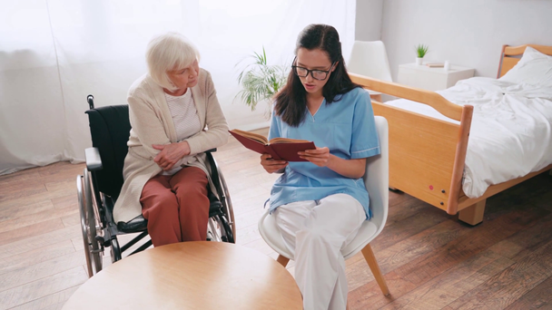jeune gériatre lecture livre à femme âgée en fauteuil roulant - Séquence, vidéo