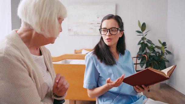 jovem enfermeira e idosa discutindo livro no hospital - Filmagem, Vídeo
