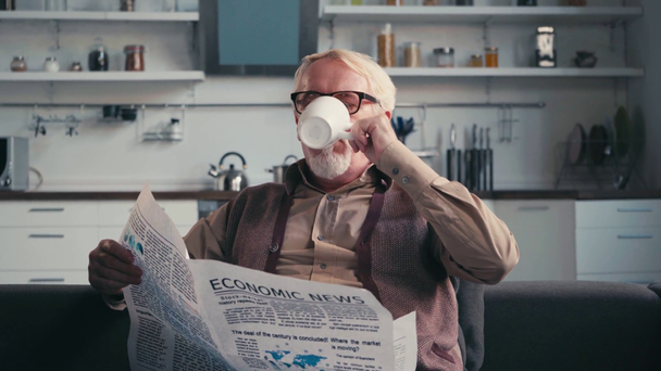 Senior man in eyeglasses reading newspaper, while drinking cup of tea at home - Felvétel, videó