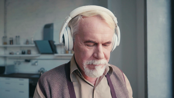 Älterer Mann mit Kopfhörern hört Musik, während er zu Hause am Fenster steht - Filmmaterial, Video