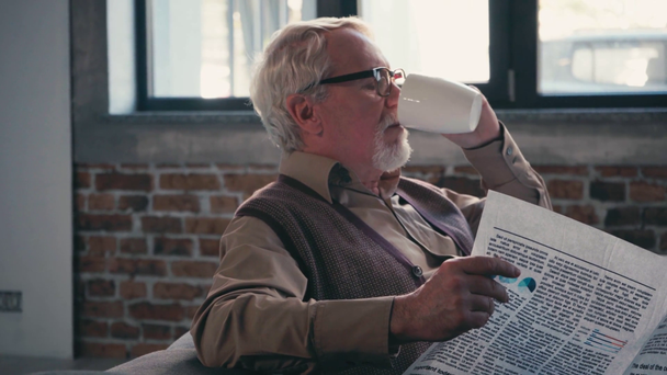Elderly man in eyeglasses reading newspaper and drinking tea at home - Footage, Video