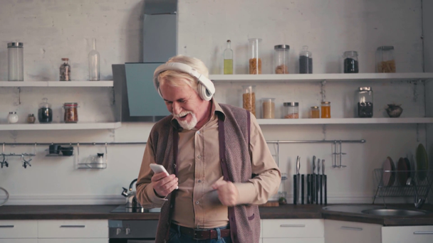 Iloinen vanhempi mies kuulokkeet älypuhelin tanssia keittiössä - Materiaali, video