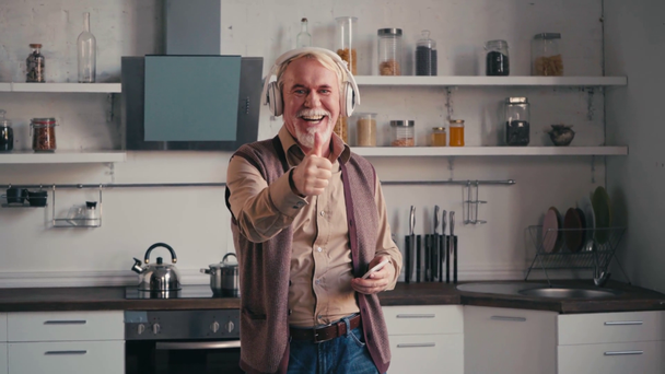 Ευτυχισμένος ηλικιωμένος με ακουστικά που χορεύει και δείχνει τον αντίχειρα του στην κουζίνα. - Πλάνα, βίντεο