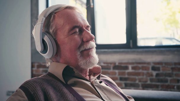 Pensionista positivo con los ojos cerrados escuchando música en auriculares en casa - Imágenes, Vídeo