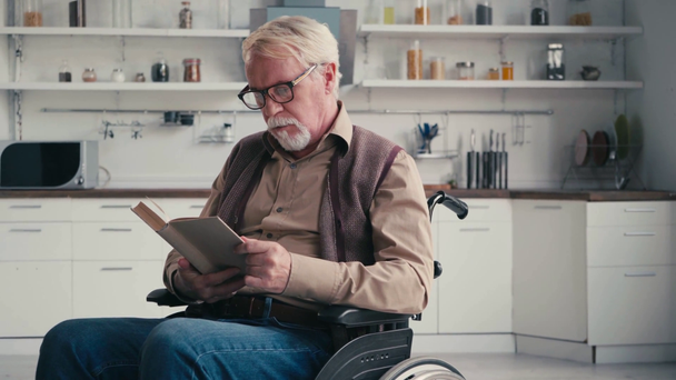 Седой мужчина в инвалидной коляске читает книгу с кухней на заднем плане - Кадры, видео