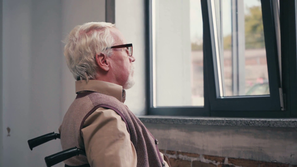Zijaanzicht van een oudere man in een rolstoel die naar binnen rijdt en thuis in het raam kijkt - Video