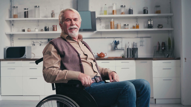 Onnellinen harmaa tukkainen mies pyörätuolissa puhumassa katsoessaan kameraa keittiössä - Materiaali, video