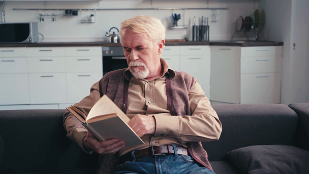 Šedovlasý muž čtení knihy, zatímco sedí na pohovce s kuchyní na pozadí - Záběry, video