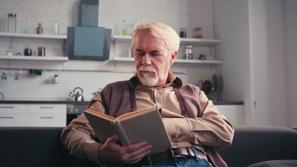 Senior άνθρωπος ανάγνωση βιβλίου, ενώ κάθεται στον καναπέ με κουζίνα στο παρασκήνιο - Πλάνα, βίντεο