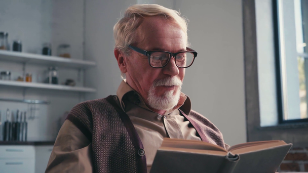 Pensionné souriant dans un livre de lecture de lunettes avec cuisine floue sur fond - Séquence, vidéo