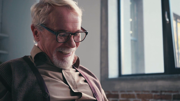 Starszy mężczyzna śmiejący się i zdejmujący okulary czytając książkę przy oknie - Materiał filmowy, wideo