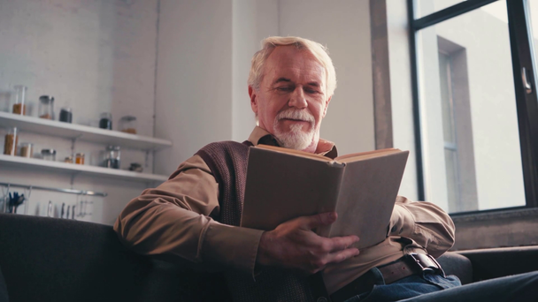 Matala kulma näkymä hymyilevä iäkäs mies lukeminen kirja istuessaan sohvalla kotona - Materiaali, video