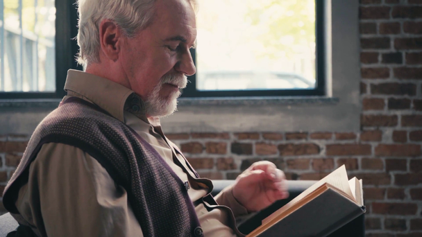 Oldalnézet pozitív idős férfi olvasás könyv és nevetés, ül az ablak mellett  - Felvétel, videó