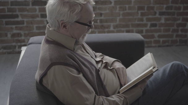 Pensionista folheando através do livro e sentado no sofá perto da parede de tijolo, monocromático - Filmagem, Vídeo