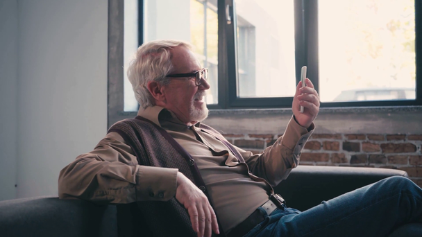 Seitenansicht des glücklichen Rentners mit winkender Hand beim Videoanruf zu Hause - Filmmaterial, Video