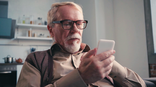 Fókuszált idős férfi szemüvegben mobiltelefont használ homályos háttérrel - Felvétel, videó