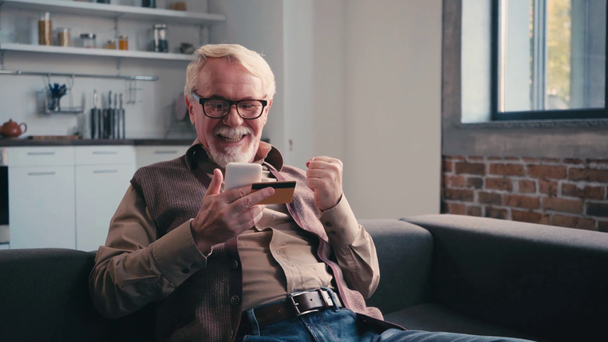 Ευτυχής συνταξιούχος που χρησιμοποιεί κινητό τηλέφωνο και πιστωτική κάρτα, ενώ κάθεται στον καναπέ στο σπίτι - Πλάνα, βίντεο