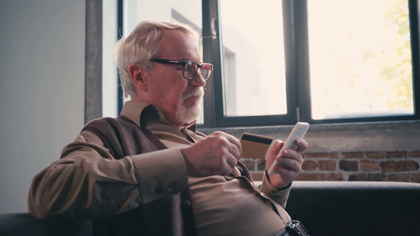 Γκρι μαλλιά άνθρωπος με πιστωτική κάρτα γραπτών μηνυμάτων στο κινητό, ενώ κάθεται κοντά στο παράθυρο - Πλάνα, βίντεο