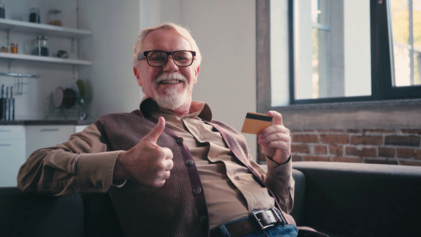 Lachende senior man toont duim omhoog en houdt credit card thuis - Video
