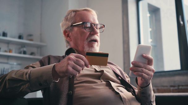 Idősebb férfi hitelkártyával SMS-ezik a mobilján, miközben otthon ül a kanapén.  - Felvétel, videó
