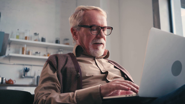 Vista basso angolo di uomo anziano digitando sul computer portatile a casa su sfondo sfocato - Filmati, video