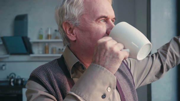 Позитивный пожилой человек смотрит в сторону, стоит у окна и пьет чай дома - Кадры, видео