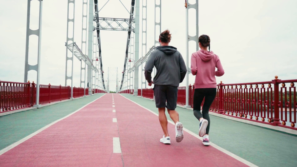 Vista trasera de pareja en ropa deportiva corriendo en puente con cielo nublado, cámara lenta - Metraje, vídeo