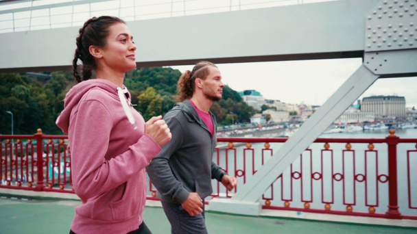 Uśmiechnięta para biegaczy biegających po moście z miastem na tle, zwolnione tempo - Materiał filmowy, wideo