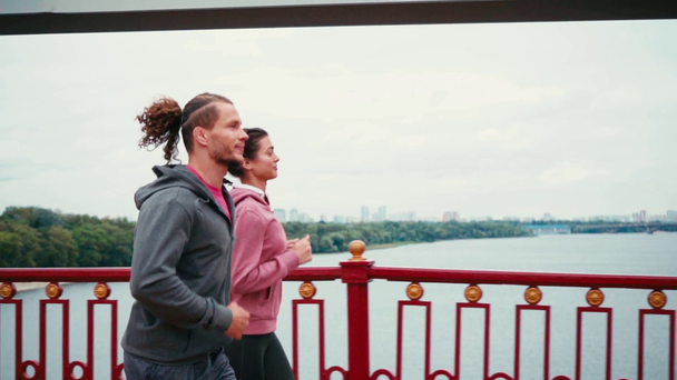 Вид сбоку пары в спортивной одежде, бегущей по мосту с природой на заднем плане - Кадры, видео