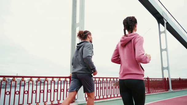 Widok wsteczny pary biegnącej wzdłuż ścieżki mostu z zachmurzonym niebem na tle - Materiał filmowy, wideo