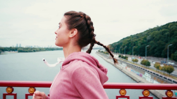 Widok z boku brunetka kobieta działa na moście z miasta na tle - Materiał filmowy, wideo