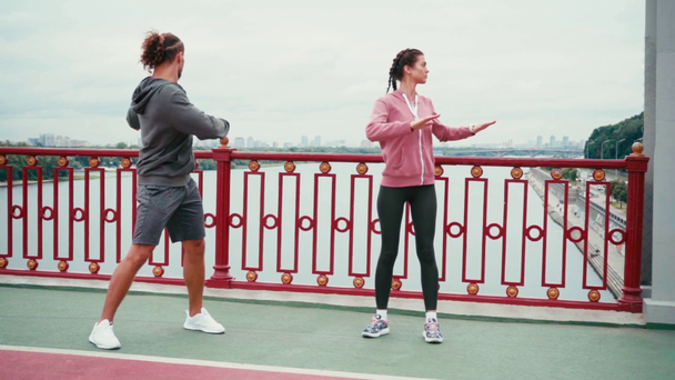 Coppia in abbigliamento sportivo che si estende sul ponte con la città sullo sfondo, slow motion - Filmati, video