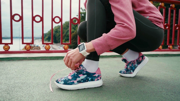 Vista recortada de la deportista con smartwatch atando cordones de zapatilla en puente - Metraje, vídeo