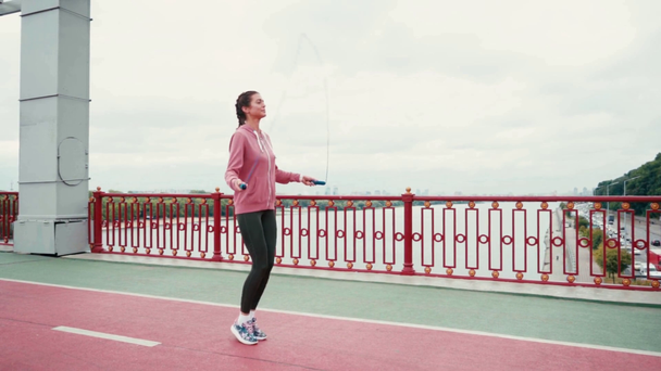 Morena deportista cuerda saltando en el puente con la ciudad en el fondo, cámara lenta - Metraje, vídeo